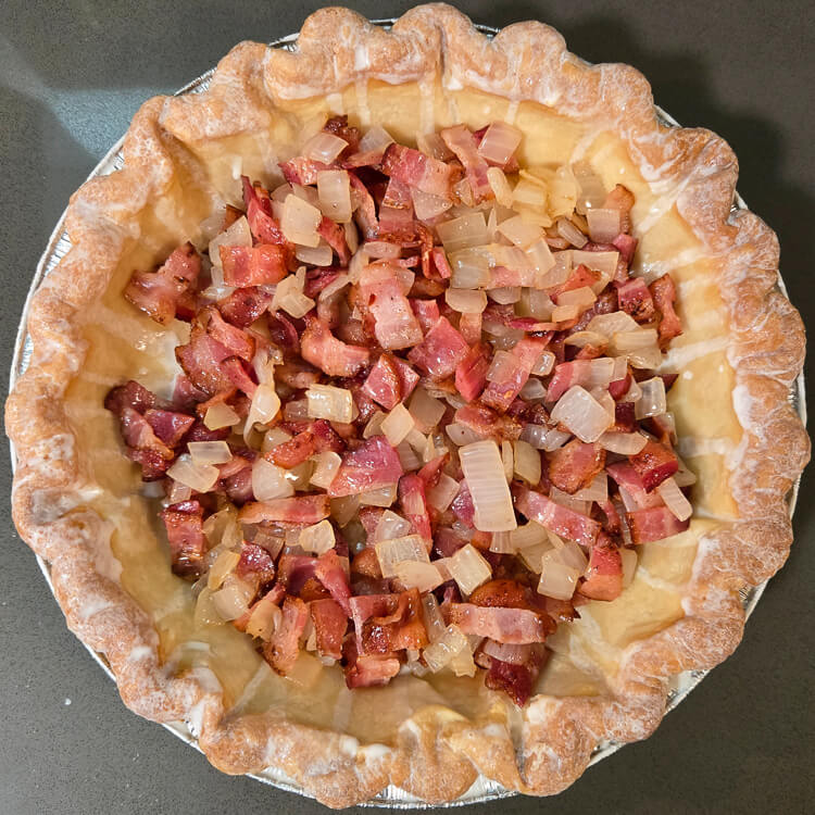 an overhead photo of a bacon and onion quiche before the egg custard is added: bacon bits and sauteed diced onion in a blind baked pie crust
