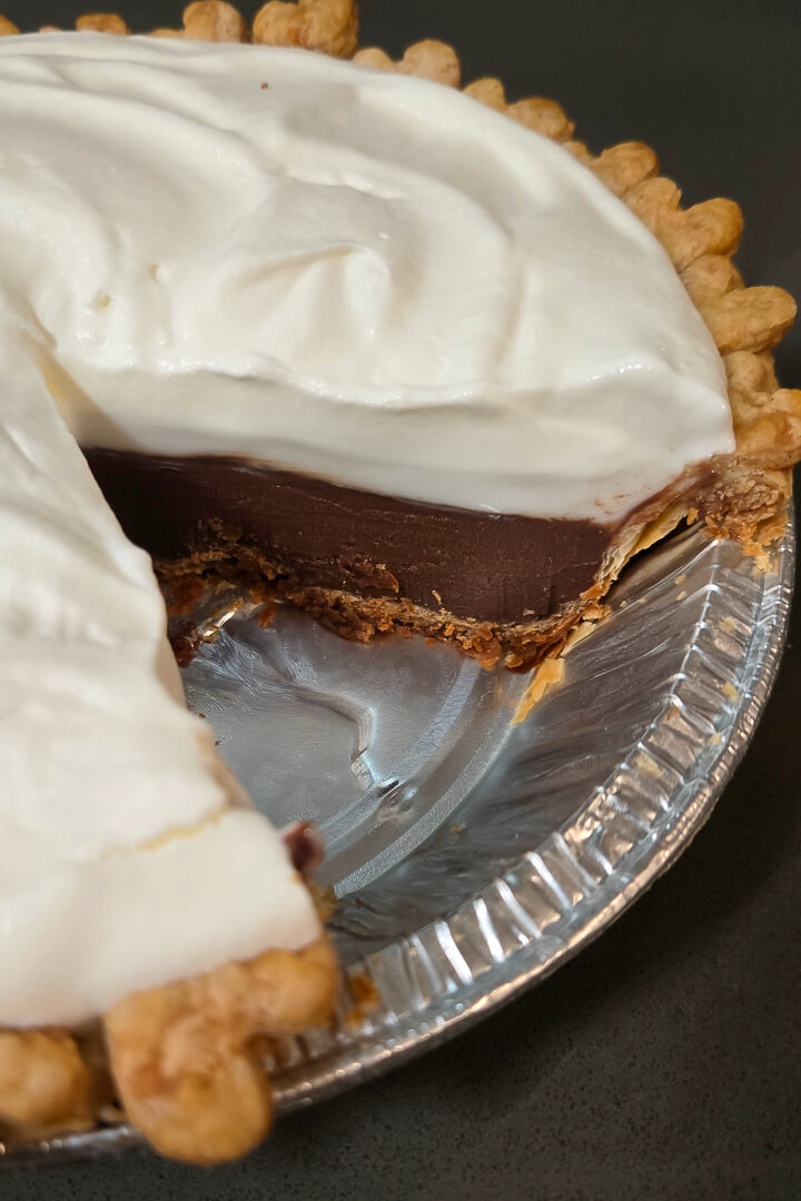close up photo of a chocolate haupia pie with a slice cut out showing a cross section with layers of golden pie crust, chocolate haupia, coconut haupia, and whipped cream