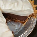 A close up of a chocolate haupia pie with a thick layer of chocolate haupia topped with fluffy whipped cream inside a flaky pie crust, shown with a slice removed to reveal the smooth, glossy chocolate filling.