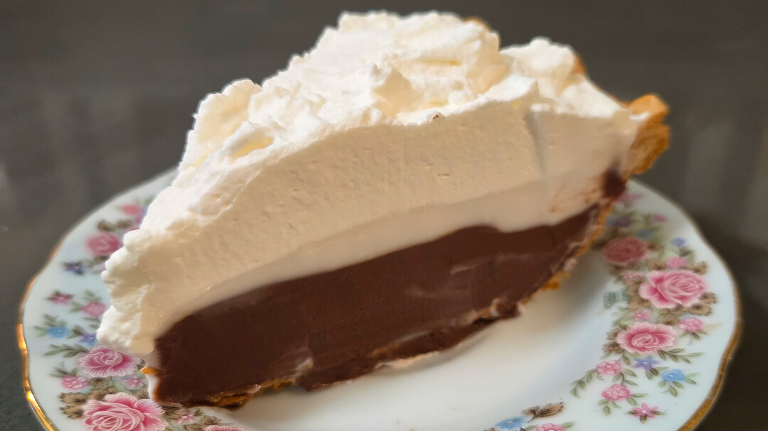 a close up photo of chocolate haupia pie with flaky pie crust, chocolate haupia, coconut haupia, and whipped cream
