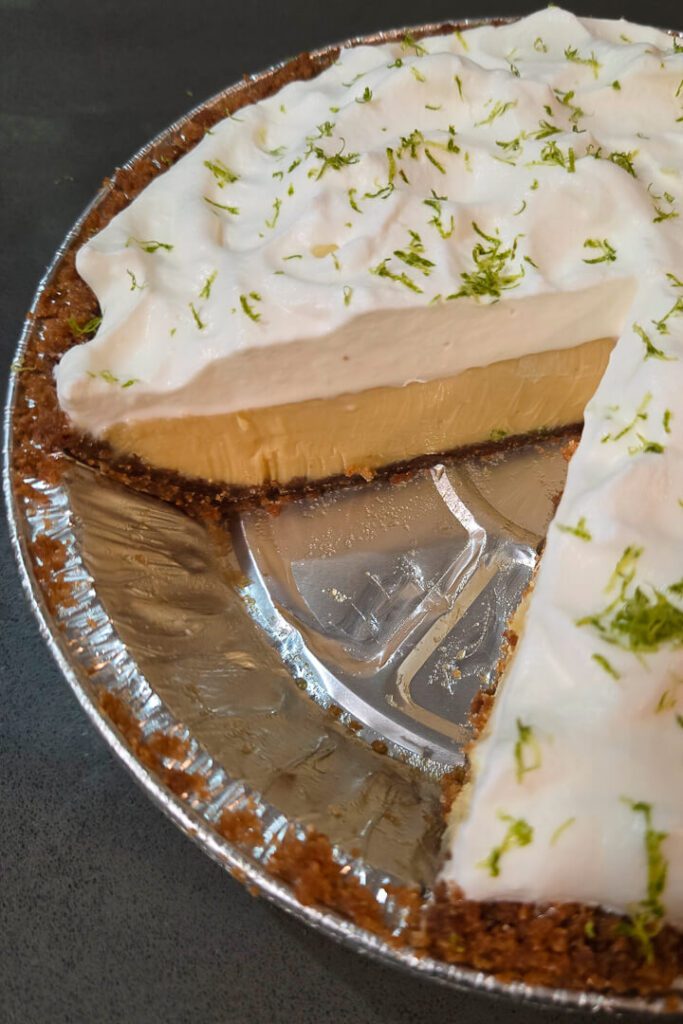 a close up photo of a key lime pie with a slice cut out so you can see the cross section with creamy lime custard, a graham cracker crust, and whipped cream topping.