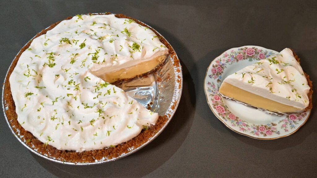 A photo of a key lime pie with a slice cut out and placed next to it on an antique floral plate