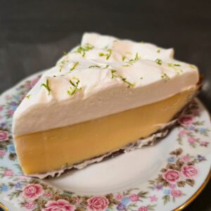 a close up photo of a slice of key lime pie on a floral plate showing graham cracker crust, smooth lime custard, and a fluffy whipped cream topping sprinkled with green lime zest
