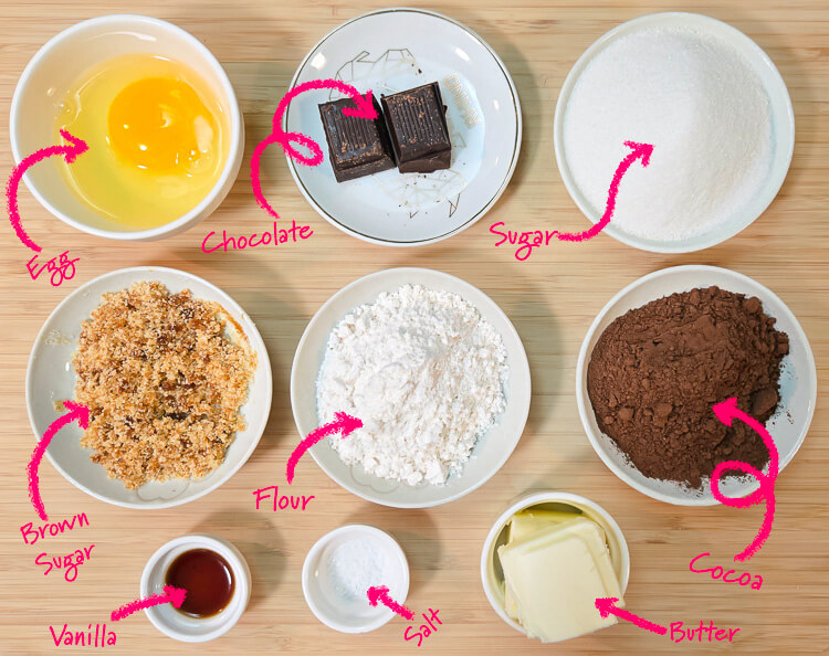 ingredients for brownie layer of a mississippi mud pie laid out on a cutting board with text labels: egg, chocolate, sugar, brown sugar, flour, cocoa, vanilla, salt, and butter