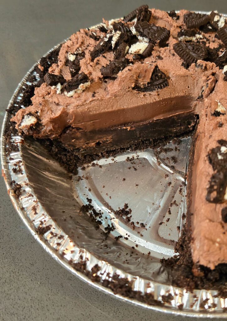a close up photo of a Mississippi mud pie with a slice cut out in an oreo crust topped with chocolate whipped cream with crushed Oreo cookies on top with a cross section showing chocolate pudding and brownies