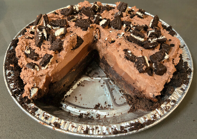 a photo of a Mississippi mud pie with a slice cut out in an oreo crust topped with chocolate whipped cream with crushed Oreo cookies on top with a cross section showing chocolate pudding and brownies