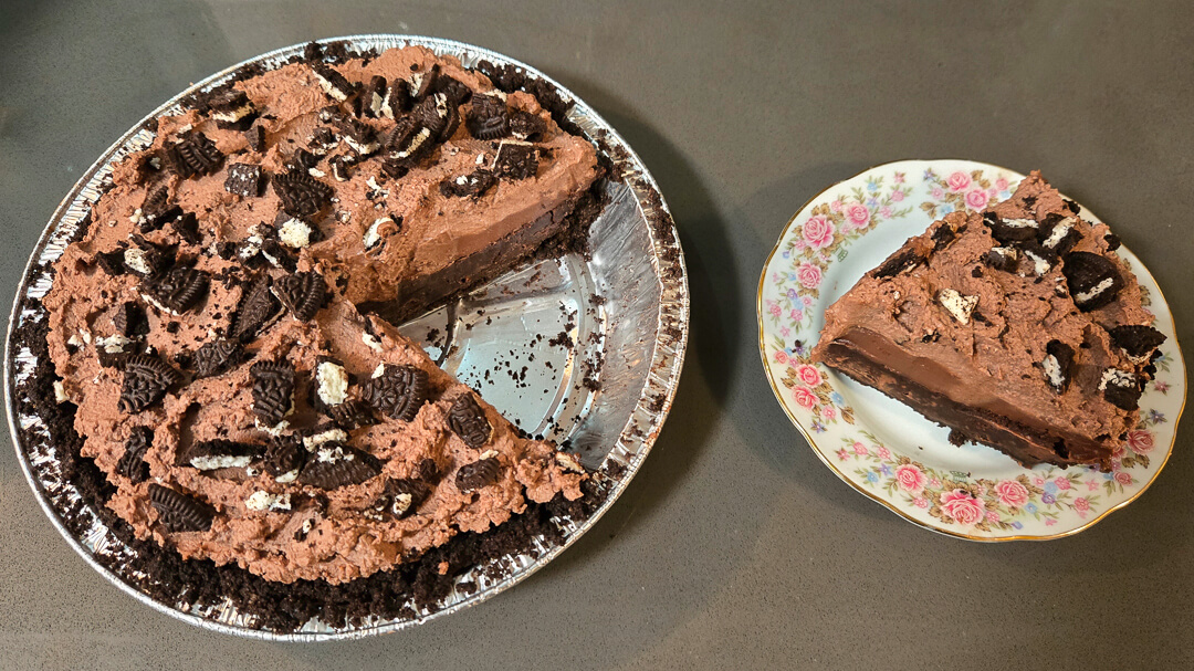a whole Mississippi Mud Pie in a metal pie tin with Oreo crust and crushed Oreo topping with a slice cut out and placed next to it on a floral antique plate