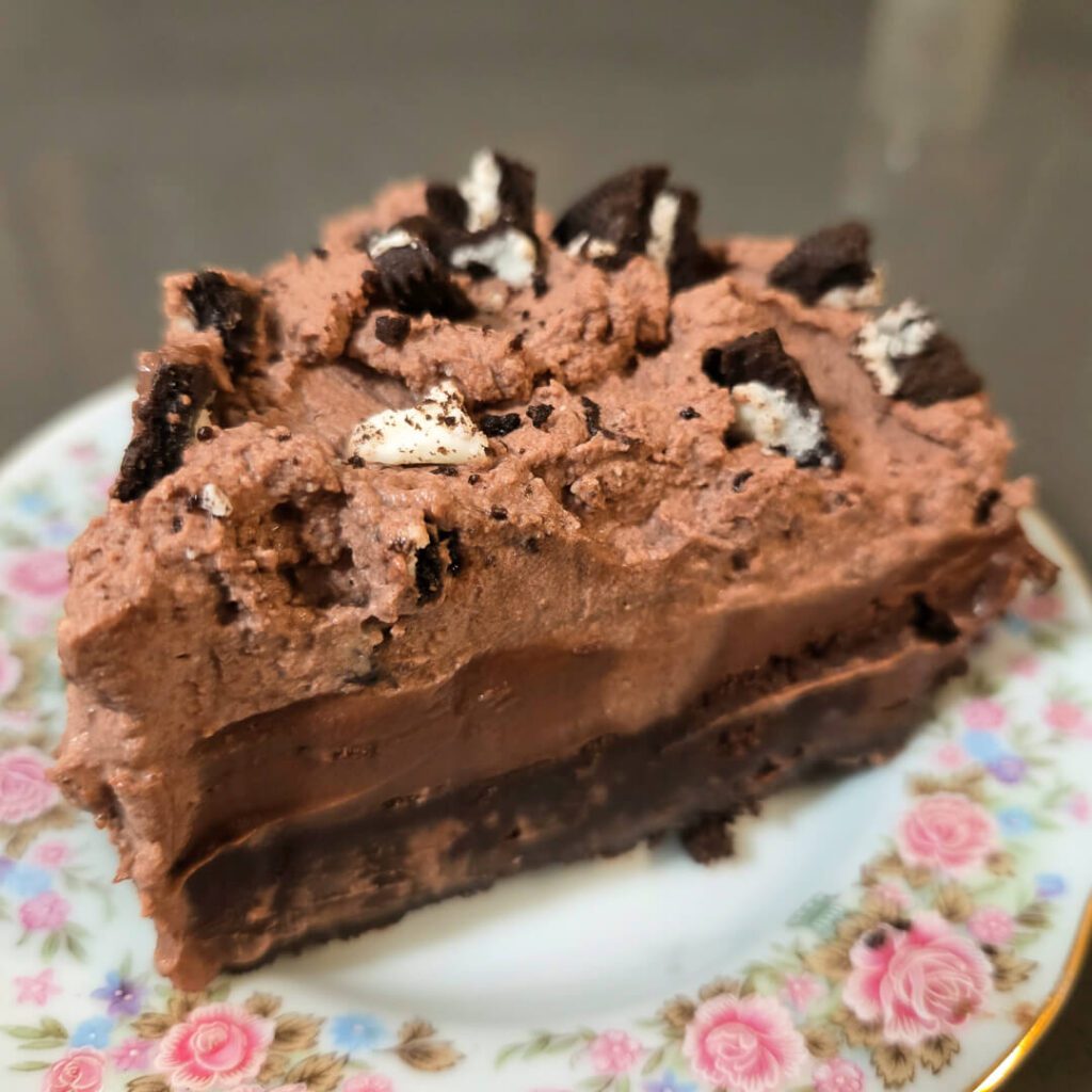 slice of mississippi mud pie on a floral antique plate showing brownies, chocolate pudding, and chocolate whipped cream topped with oreos