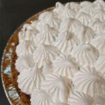 A pie topped with piped white meringue in decorative swirls, shown at an angle in a foil pie pan with a graham cracker crust.