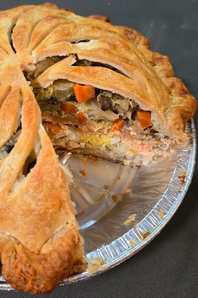 a close up photo of an Aalaskan salmon pie after a slice has been cut out showing the cross section of layers in a metal pie tin: rice, eggs, salmon, vegetables in a golden-brown flaky pie crust