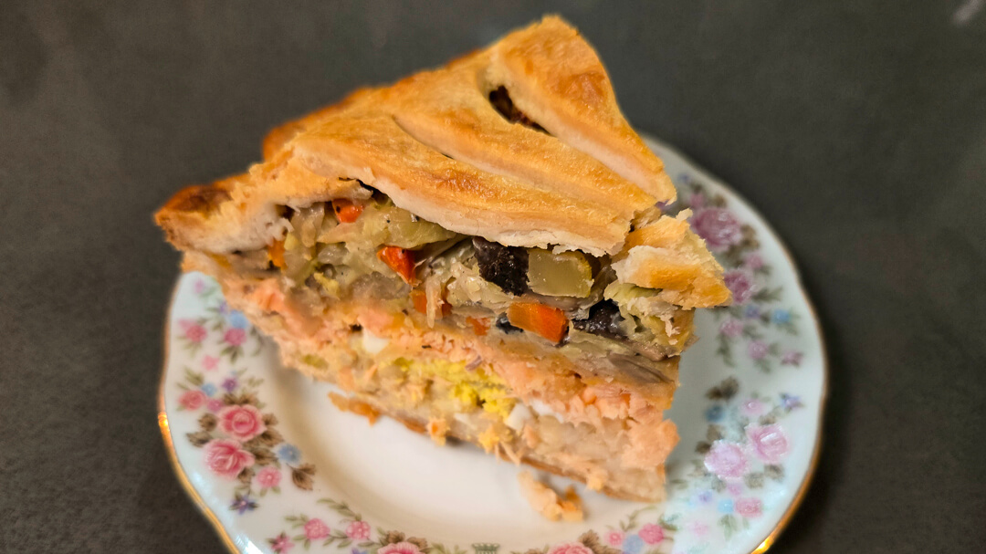 a slice of pirok (alaskan salmon pie) showing a cross section of the layers in a golden crust: brown rice, hard-boiled eggs, flaked salmon, cheese, and sauteed vegetables on an antique floral plate