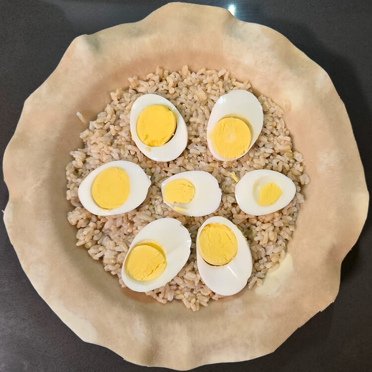 an overhead view of a savory Alaskan salmon pie (pirok) being assembled. Pie dough is fitted into a pie tin with a layer of brown rice and sliced hard-boiled eggs spread on top