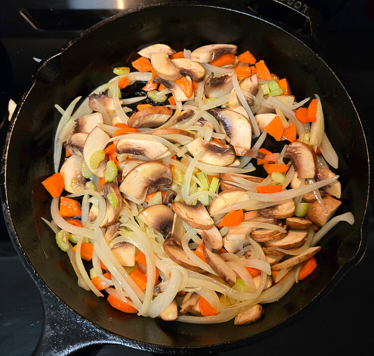 vegetables being sauteed in a cast iron pan for pirok: sliced onions, diced carrots, diced celery, and sliced mushrooms
