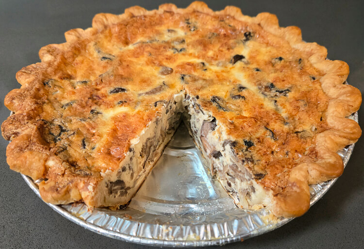a photo of a whole mushroom quiche with a slice cut out so you can see the crimped pie crust and creamy custard with mushrooms