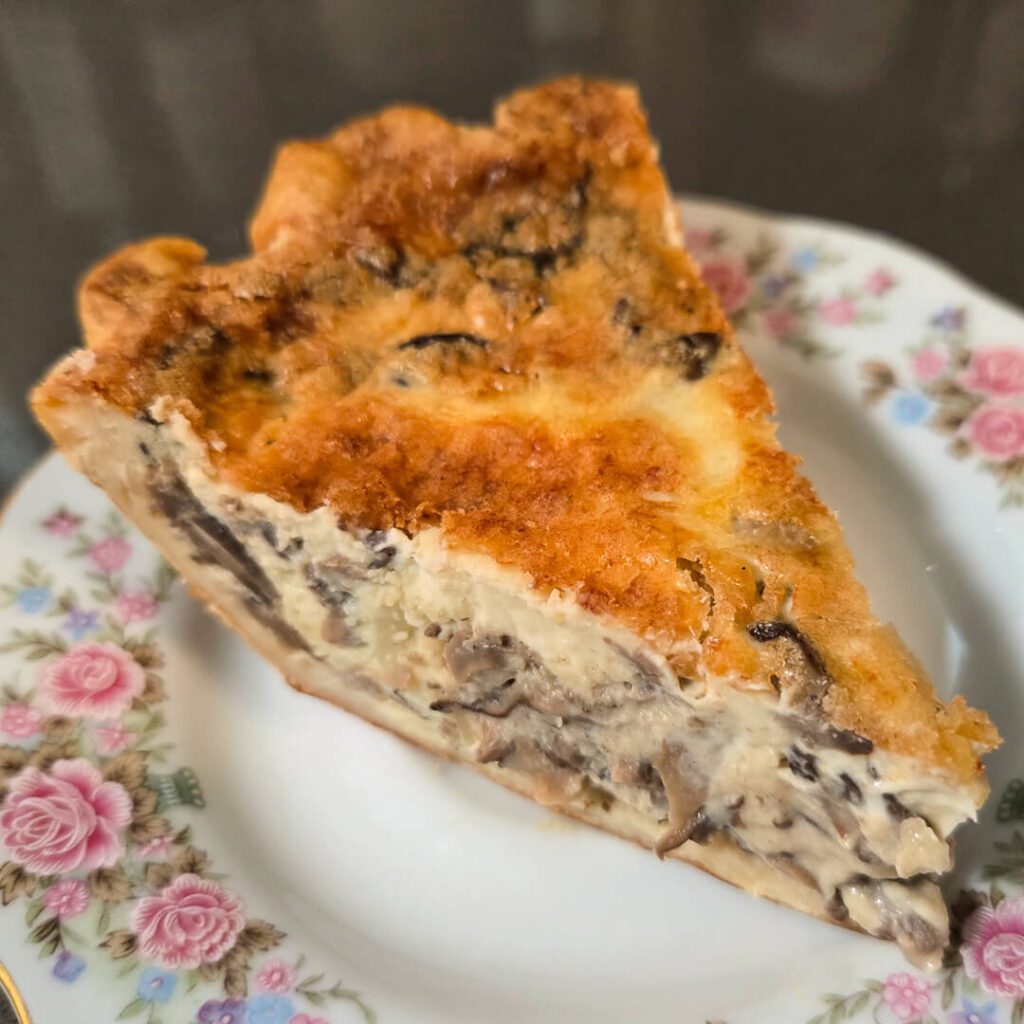 a close up overhead photo of a slice of cheesy quiche filled with mushrooms and creamy custard on a floral antique plate