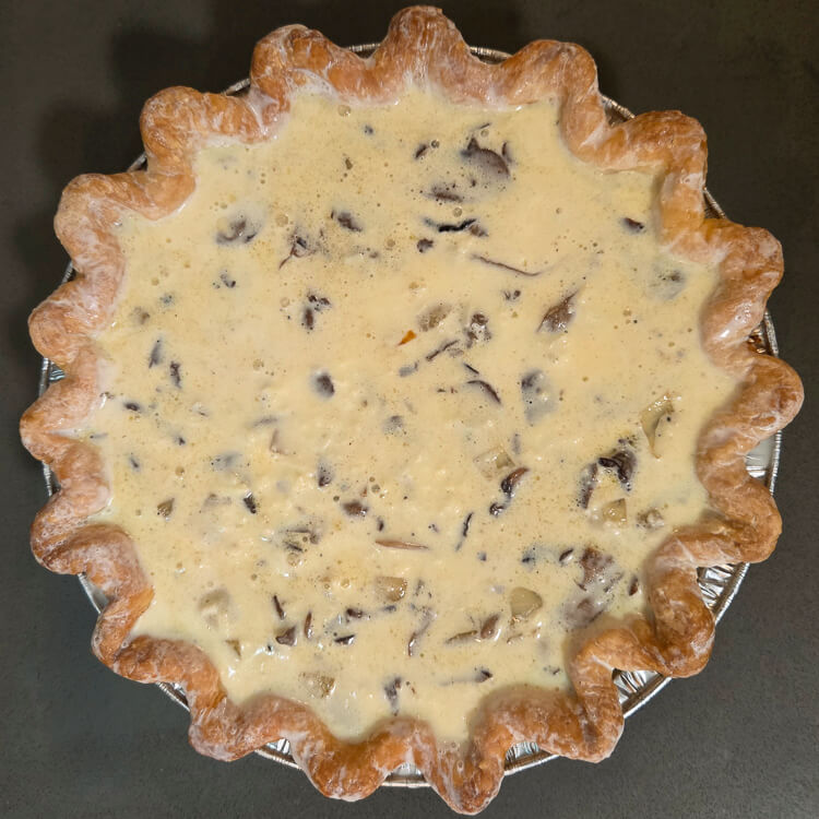 an overhead photo of an unbaked mushroom quiche with unbaked egg custard, cheese, onions, and mushrooms in a crimped pie crust with the edges brushed with cream