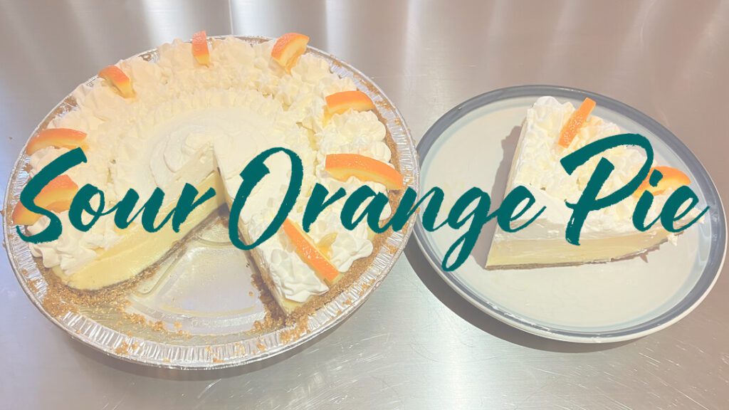 a photo of a whole sour orange pie with a slice cut out showing piped whipped cream, decorative orange slices, and creamy orange custard. It has a slice cut out and placed on a plate next to it. Text overlay that reads "Sour Orange Pie"