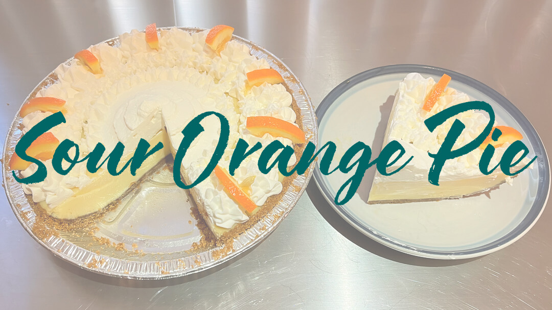 a photo of a whole sour orange pie with a slice cut out showing piped whipped cream, decorative orange slices, and creamy orange custard. It has a slice cut out and placed on a plate next to it. Text overlay that reads "Sour orange pie recipe"