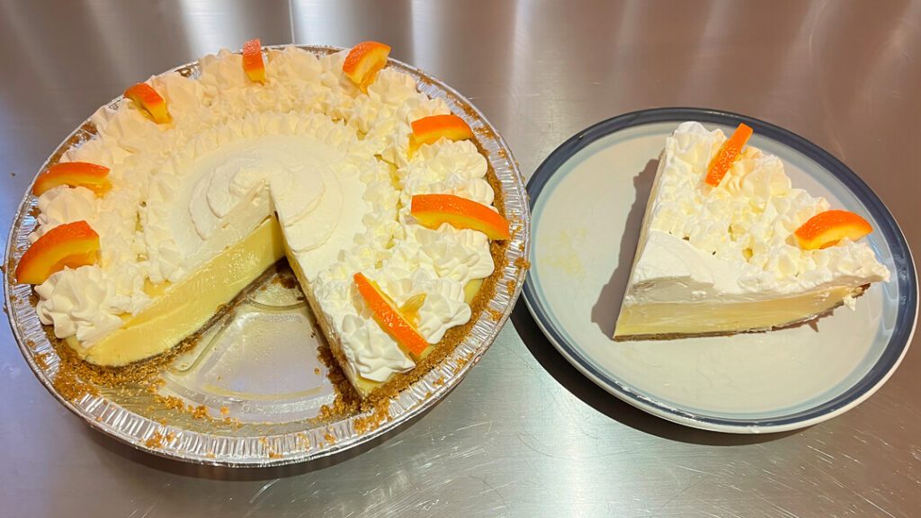 a photo of a whole sour orange pie with a slice cut out showing piped whipped cream, decorative orange slices, and creamy orange custard. It has a slice cut out and placed on a plate next to it.