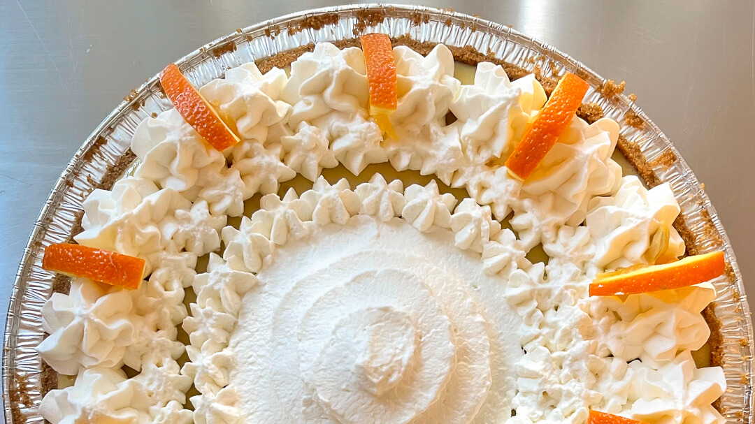 an overhead landscape photo of approximately half of a sour orange pie with piped whipped cream and decorative orange slices in a graham cracker crust