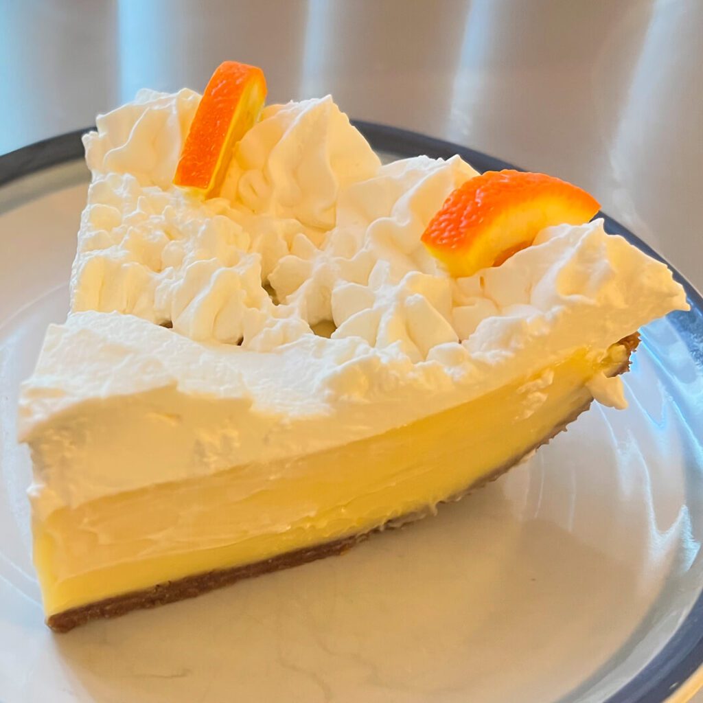 a close up photo of a slice or sour orange pie with creamy custard in a graham cracker crust with piped whipped cream on top decorated with orange slices