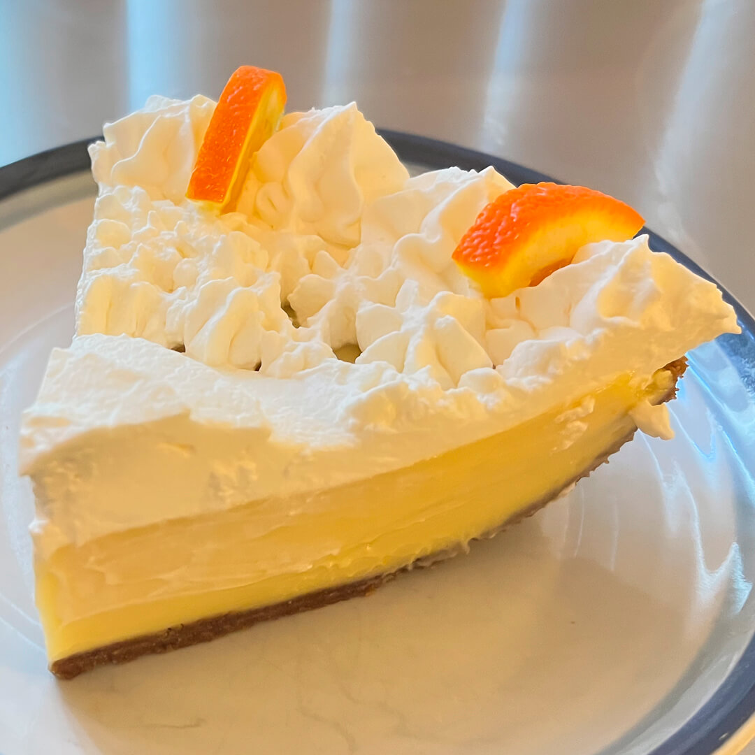 a close up photo of a slice or sour orange pie with creamy custard in a graham cracker crust with piped whipped cream on top decorated with orange slices