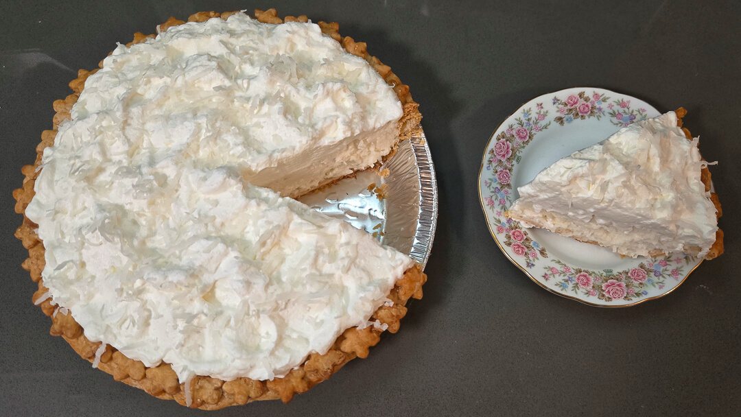 an overhead photo of a whole white christmas pie with a slice cut out and placed next to it on a plate. The pie is a chiffon pie with a fluffy whipped cream topping and shredded coconut making it look like snow.