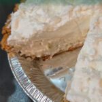 White Christmas chiffon pie in a foil pie pan with one slice missing, showing the fluffy white filling topped with whipped cream and shredded coconut.