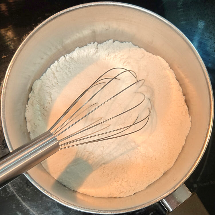 flour and sugar being whisked together in a metal pot before dissolving it in milk