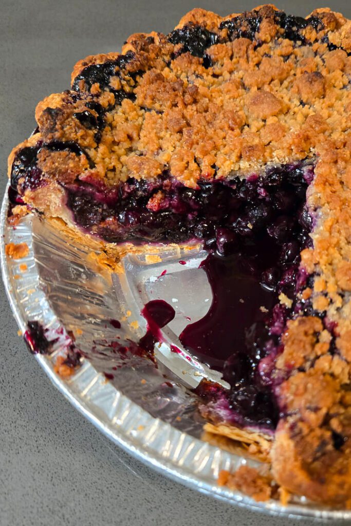 a close up photo of a blueberry pie topped with golden crumble with a slice cut out so you can see the juicy purple blueberry filling