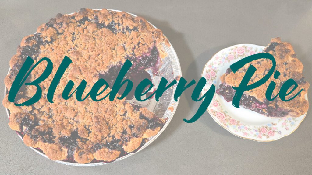 an overhead photo of a blueberry pie with crumble topping with a slice cut out and placed next to it on an antique plate with flowers. Text overlay that reads "blueberry pie"