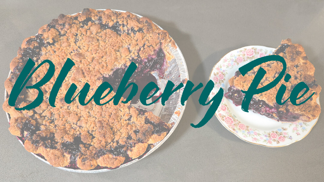 an overhead photo of a Blueberry pie recipe with crumble topping with a slice cut out and placed next to it on an antique plate with flowers. Text overlay that reads "blueberry pie"