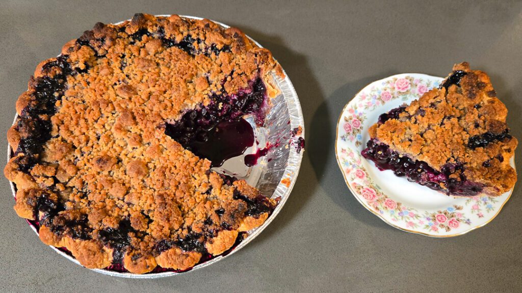 an overhead photo of a blueberry pie with crumble topping with a slice cut out and placed next to it on an antique plate with flowers