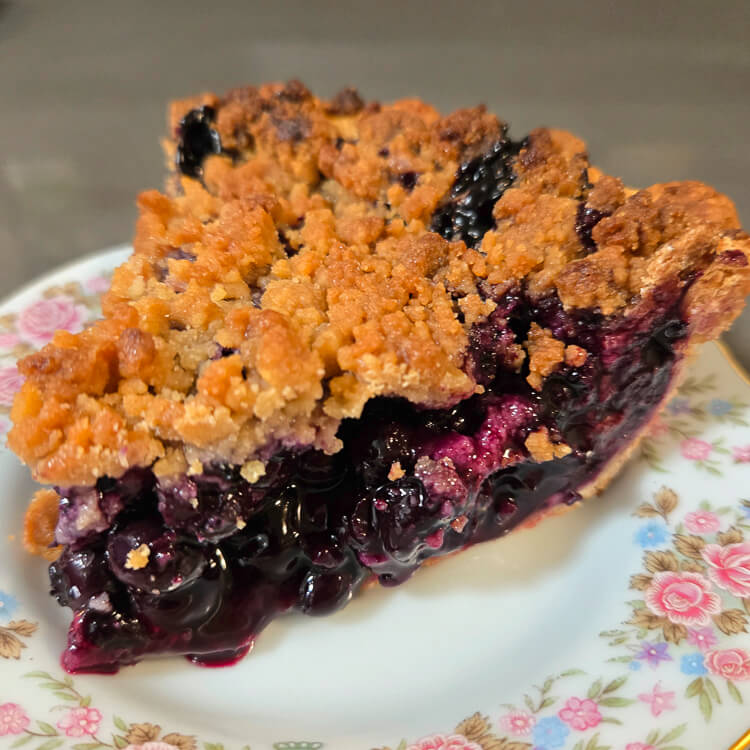Classic Blueberry Pie Recipe a close up photo of a slice of blueberry pie with glossy blueberry filling in a golden crust topped with golden crumble on an antique plate with flowers