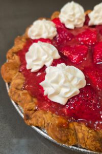 a close up photo of fresh strawberry pie with glossy red glaze topped with piped whipped cream in a crimped pie crust