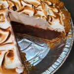 A chocolate meringue pie in a flaky pie crust with a slice removed, showing a thick layer of dark chocolate pudding filling topped with billowy golden-toasted meringue.