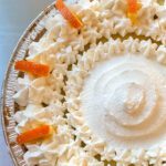 An overhead close-up of a sour orange pie in a graham cracker crust, topped with piped whipped cream rosettes and decorative candied orange peel strips, with text overlay reading "Sour Orange Pie."