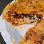 A spaghetti pie on a white plate with a slice removed, showing distinct layers of golden spaghetti parmesan crust, creamy herbed ricotta filling, and rich meat sauce topped with melted browned cheese.