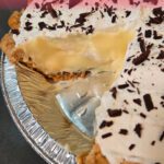 A banana cream pie in a pie tin with a slice cut out. Homemade vanilla pudding and sliced bananas make up the filling. The crust is a crimped all-butter pie crust. And whipped cream with chocolate shavings top the pie. Text overlay reads "The Best Banana Cream Pie" with the Doggone Baking logo in the bottom left corner