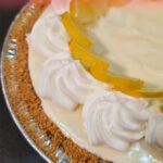 Close-up angled overhead view of a Frozen Lemonade Pie with a smooth pale yellow no-churn lemon ice cream filling, piped whipped cream rosettes around the edge, and fresh lemon slice garnishes in a graham cracker crust, with a pink and red text banner at the top reading "Frozen Lemonade Pie — with Easy No-Churn Ice Cream!" and the Doggone Baking logo in the bottom left corner.