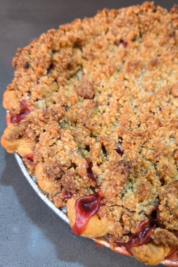 a close up photo of a strawberry rhubarb pie with crumb topping with pink filling bubbling out of a crimped all-butter pie crust