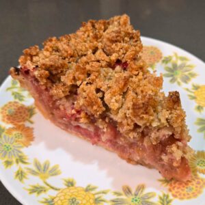 close up photo of a slice of strawberry rhubarb pie with a golden crumb topping with pink filling visible on a white floral plate