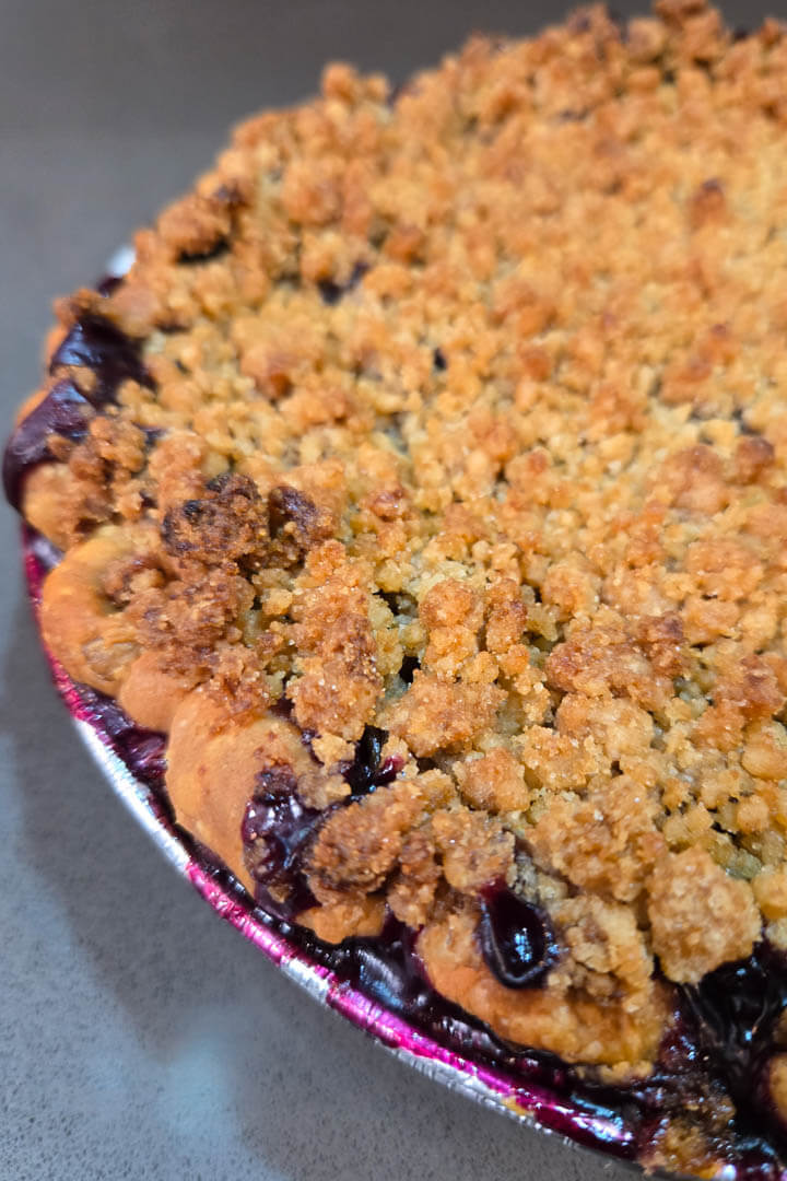 blueberry rhubarb pie, also called bluebarb pie. Close up photo showing crimped pie crust with crunchy crumble topping with dark purple blueberry rhubarb pie filling bubbling out