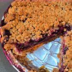 A Pinterest pin for a blueberry rhubarb pie recipe. The image shows a top-down angled view of a blueberry rhubarb pie with a thick golden crumb topping and deep purple blueberry rhubarb filling visible where a slice has been removed, with the Doggone Baking logo at the bottom. Text overlay reads "Blueberry Rhubarb Pie | 'Bluebarb Pie'."