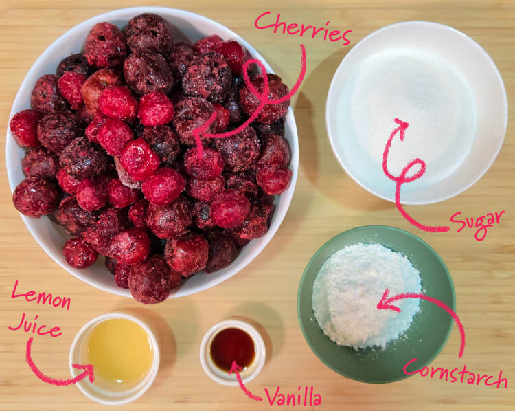 an overhead view of ingredients for homemade cherry pie filling laid out on a cutting board with text labels: cherries, sugar, lemon juice, vanilla, cornstarch