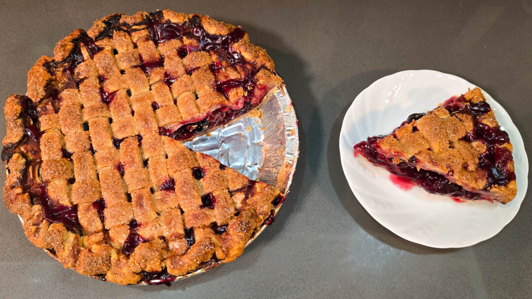 an overhead photo of a cherry pie with a lattice top with a slice cut out and placed next to it on a white plate. 