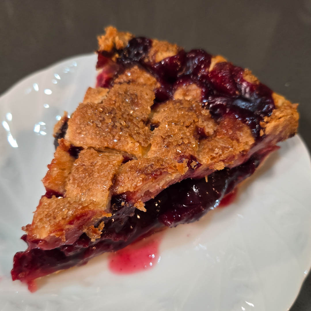 a photo of a slice of classic cherry pie with juicy red filling and a golden flaky all-butter pie crust shaped into a lattice