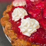 Angled overhead close-up of a fresh strawberry pie with glossy red strawberry glaze and three piped whipped cream rosettes in a crimped pastry crust, with a pink and red text banner at the top reading "Mother's Day Brunch Ideas — Strawberry Pie" and the Doggone Baking logo in the bottom left corner.