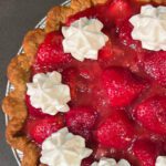Overhead view of a fresh strawberry pie filled with whole glazed strawberries and piped whipped cream rosettes in a golden crimped pastry crust. Text overlay reads Strawberry Pie Fresh Strawberries in Homemade Glaze.