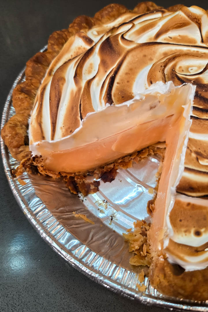 grapefruit meringue pie close up photo with a slice cut out showing a cross section of flaky pie crust, pale pink grapefruit curd, and a toasted fluffy meringue topping
